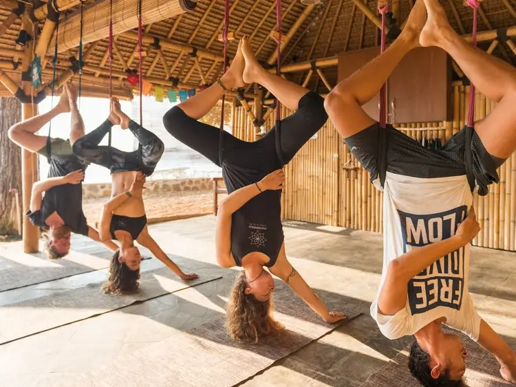 Un grupo en columpios de yoga aéreo, boca abajo bajo un techo de bambú junto a la playa