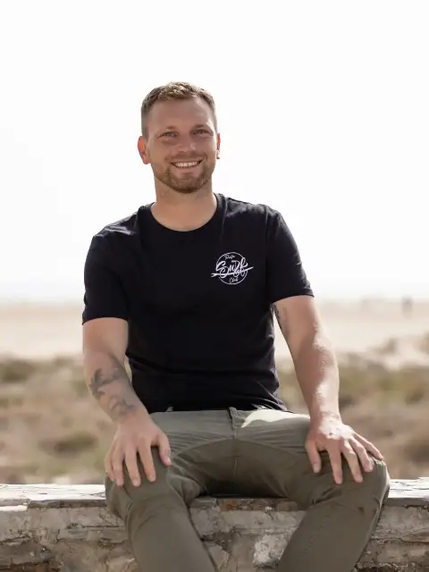 Mark, de Tarifa Surf Club, sentado en un muro en un acceso a la playa, con arena y dunas al fondo