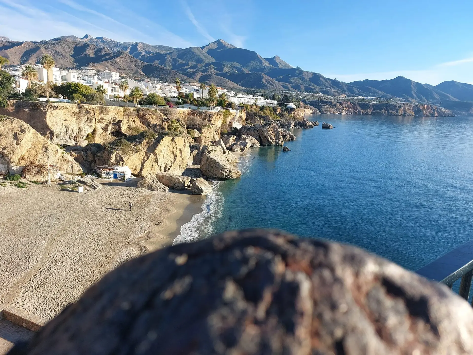 Nerja en invierno: casas blancas sobre una bahía en calma con las montañas de la Almijara al fondo y una playa vacía bajo un sol bajo y brillante